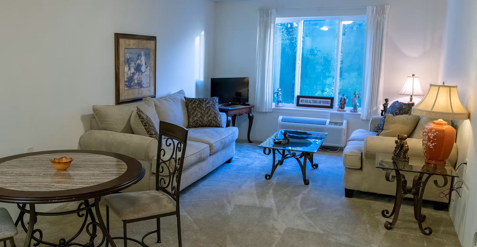 A cozy living room with two beige sofas facing each other, a glass coffee table in the center, a small TV on a wooden side table near a window with white curtains, and a round dining table with two chairs in the foreground. The room is softly lit by a table lamp with an orange base and decorated with figurines and a framed picture on the wall.