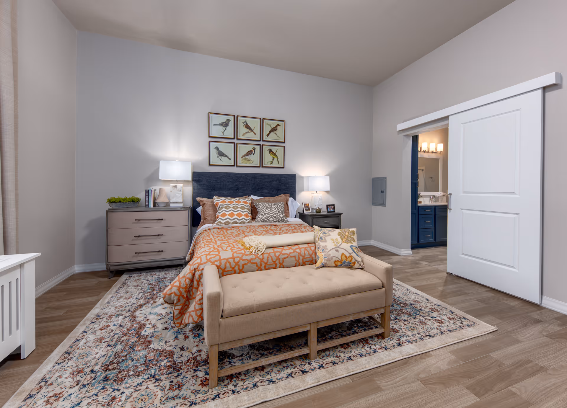 Well-lit furnished bedroom with a bed, upholstered bench, nightstands and lamps, bird prints above the headboard, and a sliding barn door to an en-suite bathroom.