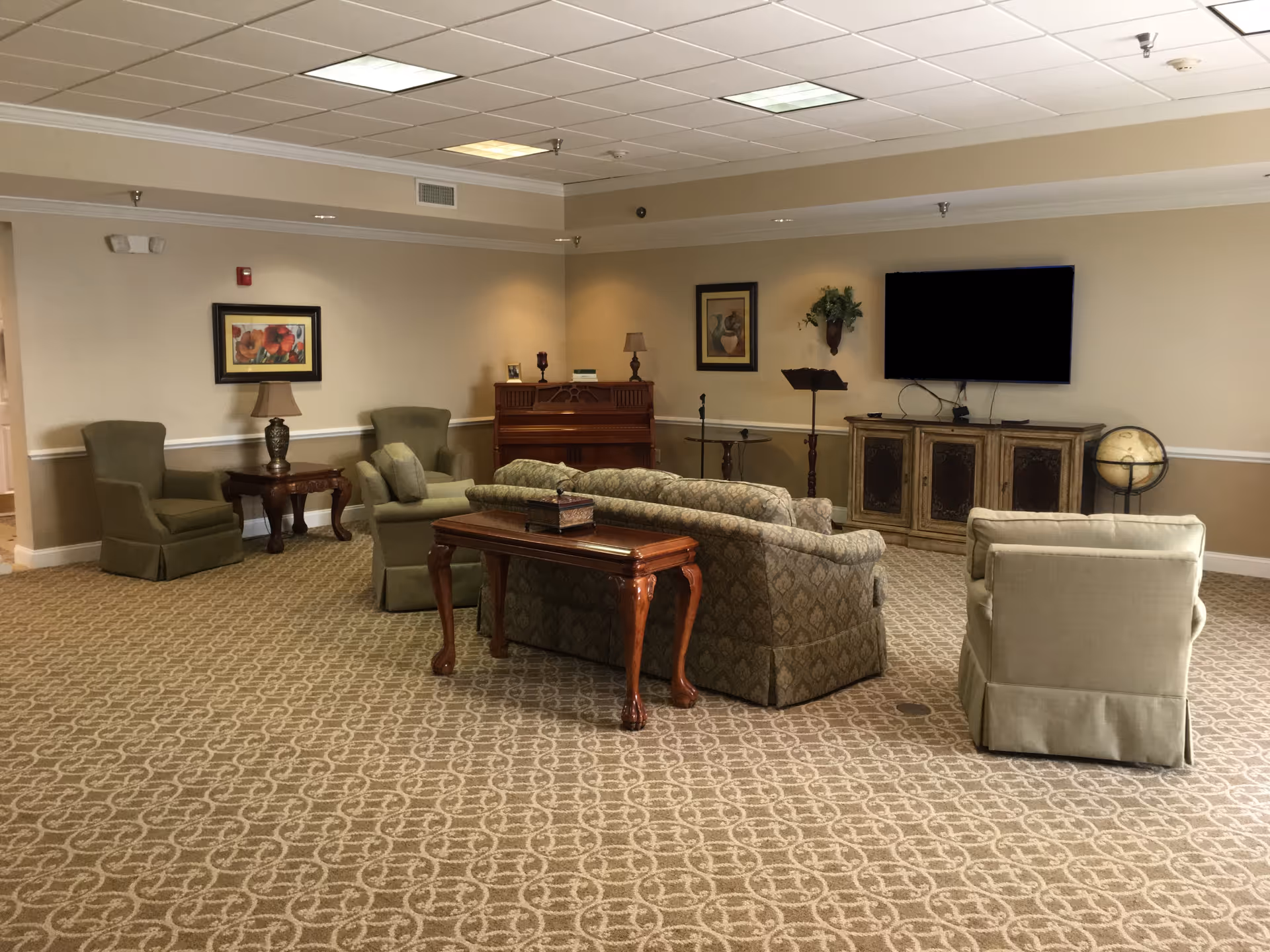 A spacious living room area with patterned carpet, beige walls, and a white ceiling. The room features a patterned sofa, two green armchairs, a beige armchair, a wooden side table with a lamp, a wooden console table behind the sofa, a piano against the wall, a flat-screen TV mounted on the wall above a decorative cabinet, a globe on a stand, and framed artwork on the walls.