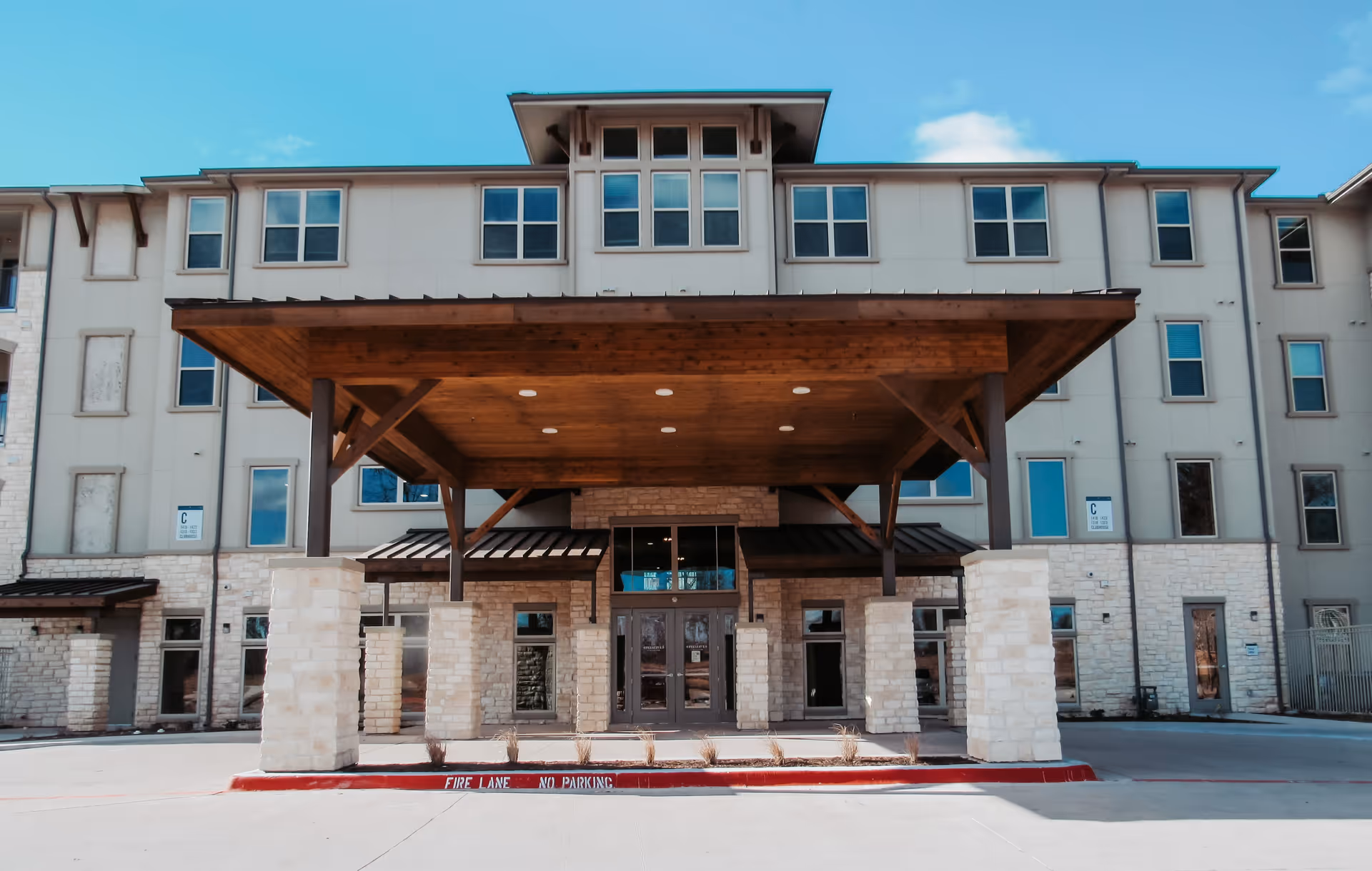 Front entrance of a multi-story senior living building with a large wooden covered porte-cochère supported by stone columns.