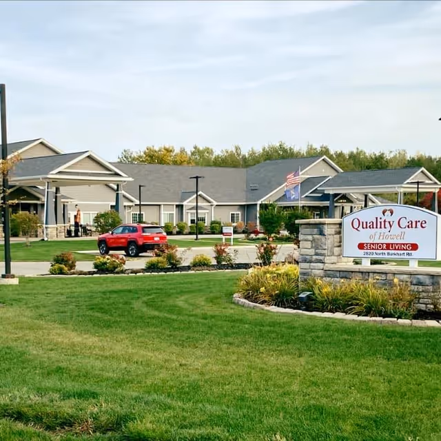 Exterior view of Quality Care of Howell senior living facility with a well-maintained lawn, a red car parked near the entrance, and a stone sign displaying the facility's name and address.