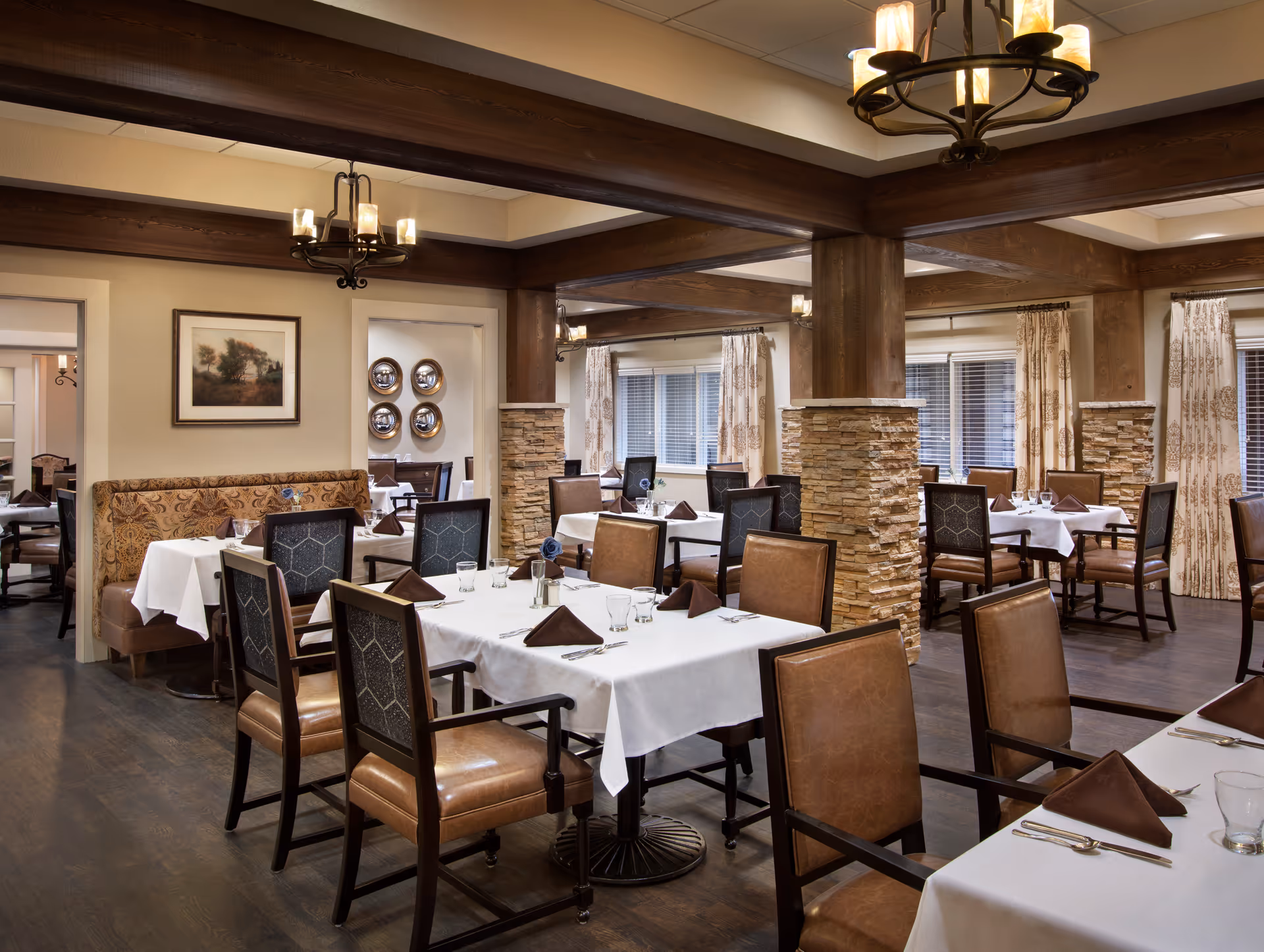 A well-lit dining room with multiple tables covered in white tablecloths and set with brown napkins, glasses, and silverware. The room features wooden beams and stone pillars, with upholstered chairs and patterned curtains on the windows. Wall decorations include framed artwork and decorative plates.