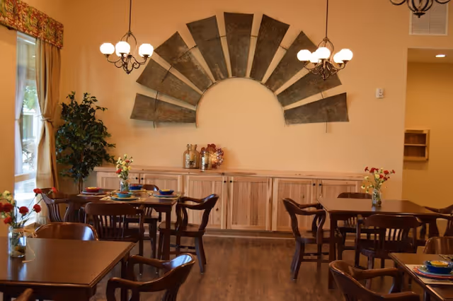 A dining room with wooden tables and chairs, each table decorated with small flower vases and place settings. The room has warm lighting from chandeliers and natural light coming through a window with curtains. A large decorative metal wall art piece shaped like a fan is mounted on the wall above a wooden cabinet.