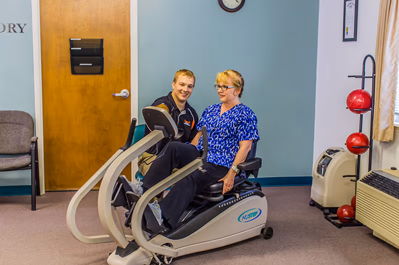A woman is seated on a recumbent exercise bike in a therapy room, assisted by a male therapist who is smiling and providing support. The room has a wooden door, chairs, a rack with red exercise balls, and medical equipment.