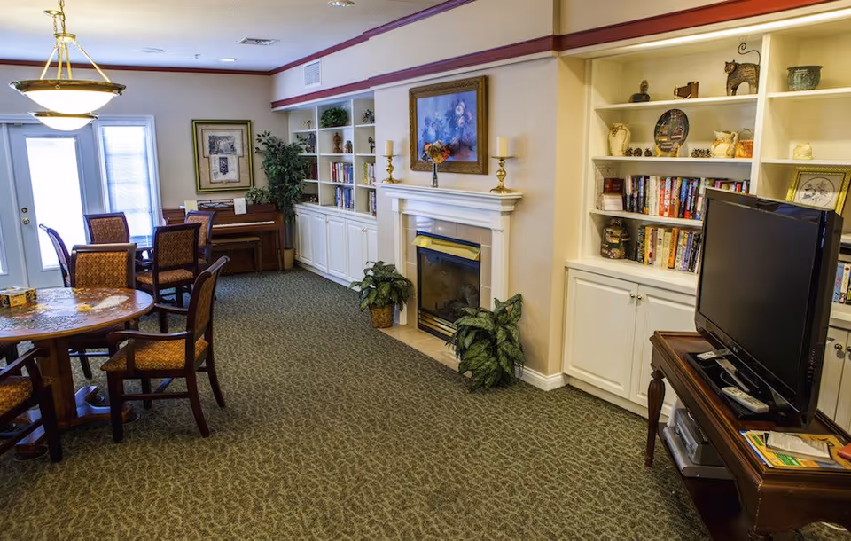 Cozy senior living common room with round dining tables and chairs, built-in bookshelves, a fireplace, and a television.