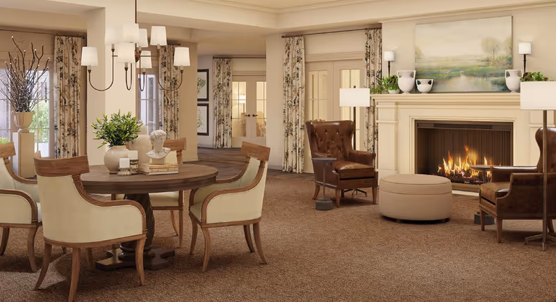 A cozy senior living common area featuring a round wooden table with four upholstered chairs, a decorative bust, and a vase with greenery. In the background, there are large windows with floral curtains, a fireplace with a fire burning, two brown leather armchairs, a round ottoman, and framed artwork above the mantel.