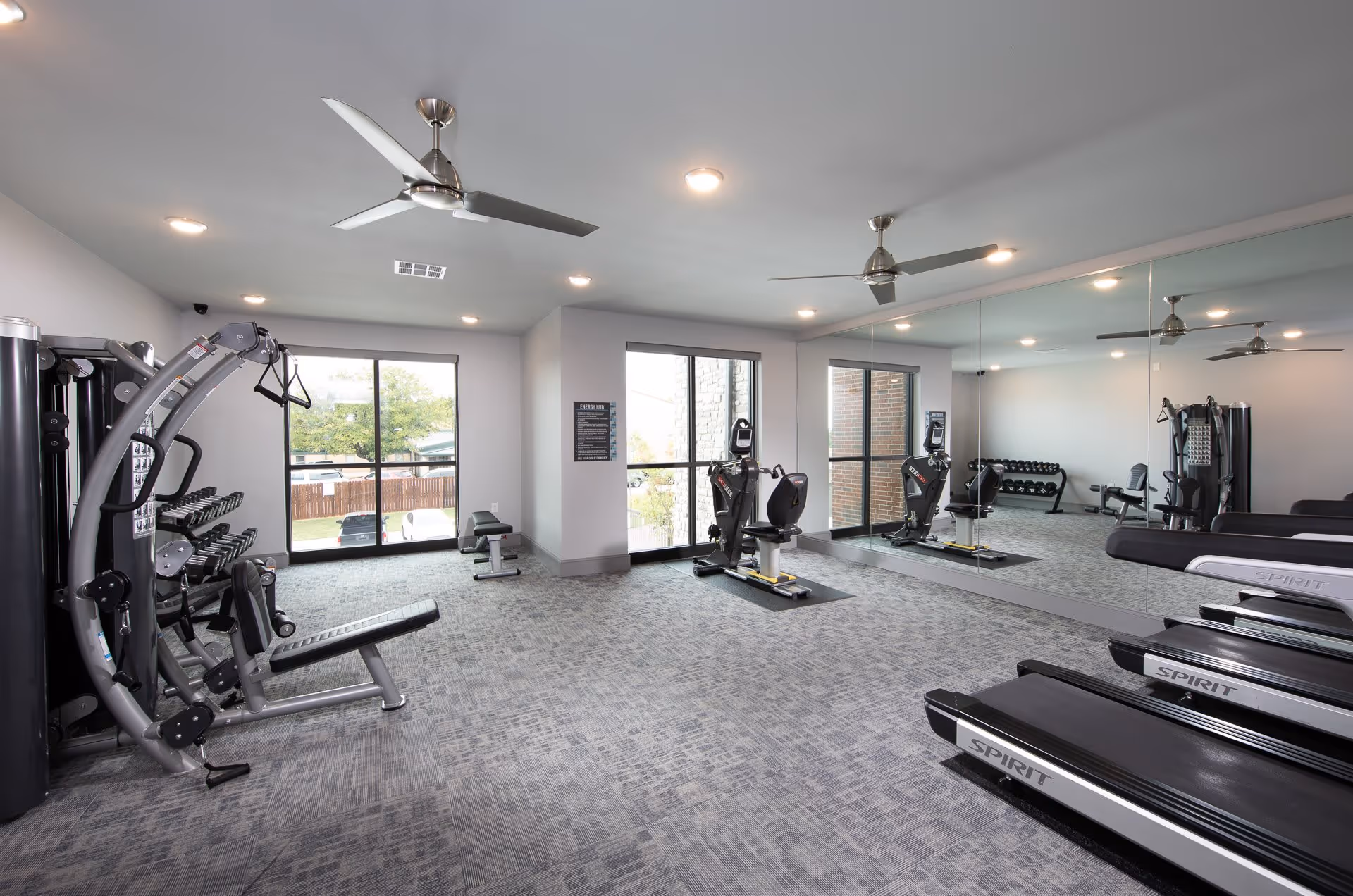A modern fitness room with exercise equipment including treadmills, a multi-function weight machine, dumbbells on a rack, and a recumbent exercise bike. The room has large windows letting in natural light, a wall covered with mirrors, ceiling fans, and recessed lighting.