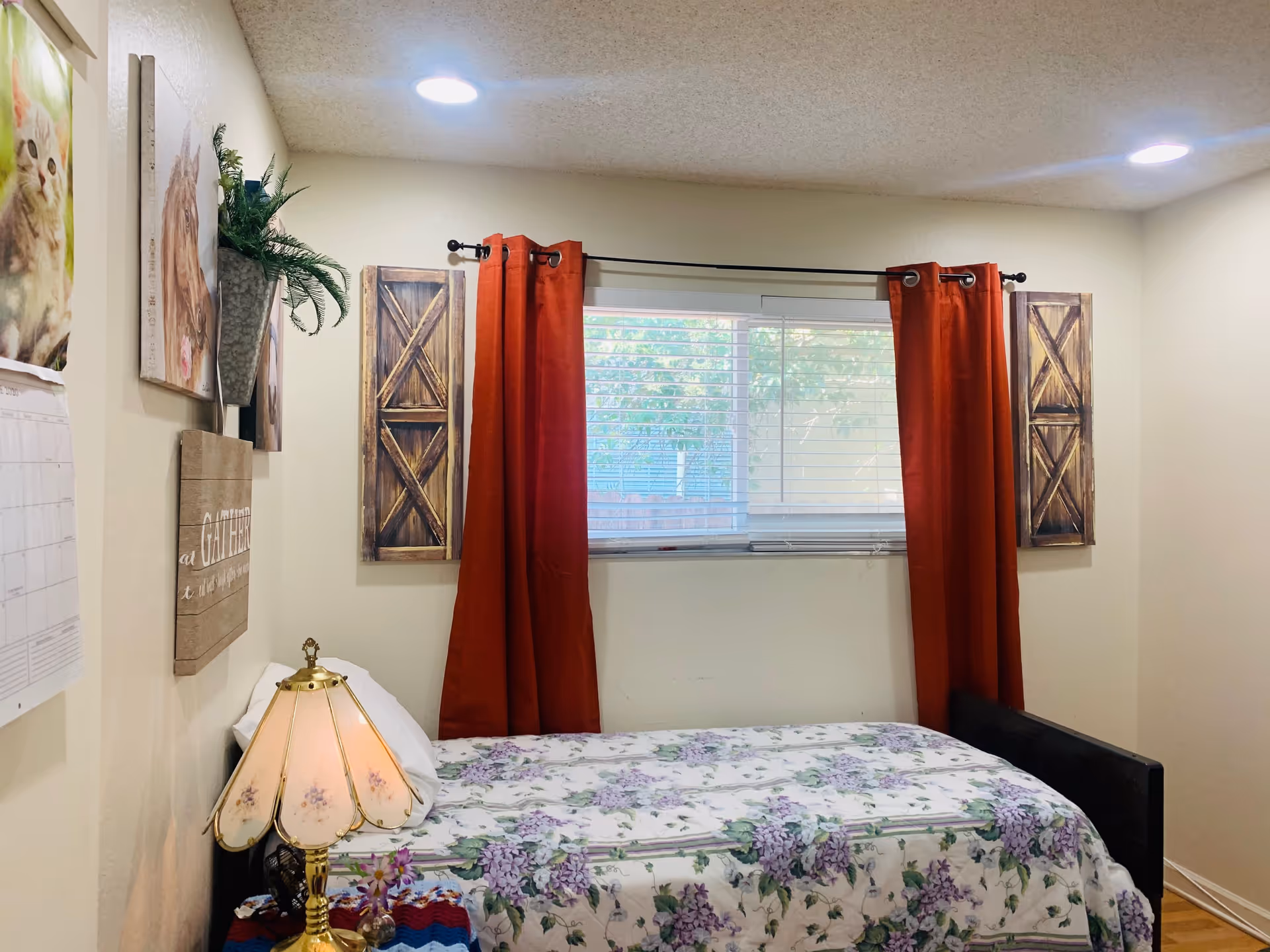 A small bedroom with a single bed covered in a floral bedspread. The bed is positioned against a wall with a window above it, framed by red curtains and wooden decorative panels on each side. On the left wall, there are pictures of animals and a wall planter with greenery. A vintage-style table lamp with a floral design is on a small table next to the bed. The room has a cozy and simple decor with soft lighting from ceiling lights.