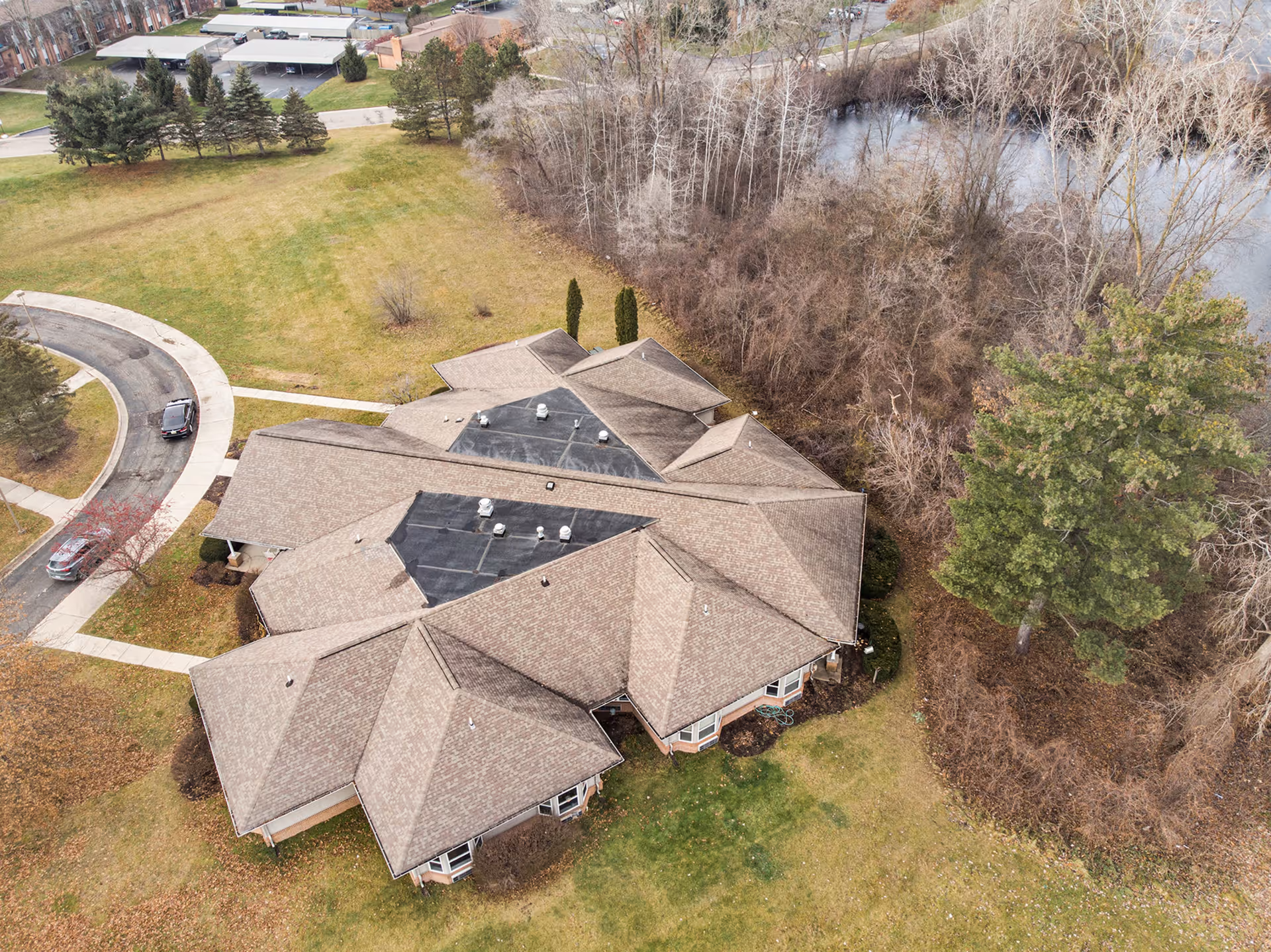 Aerial view of a single-story building with a complex roof structure surrounded by grass, trees, and a curved driveway with two parked cars. There is a wooded area and a body of water visible in the background.