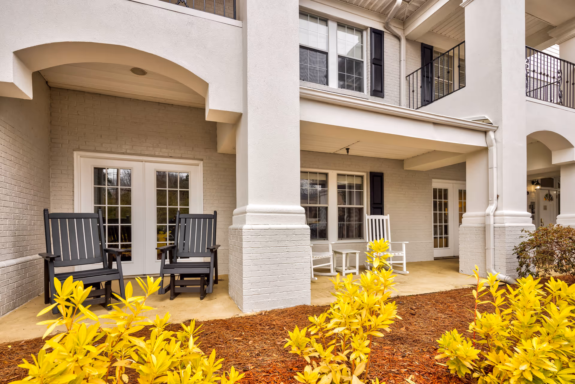 Outdoor covered patio area of a senior living facility with black and white rocking chairs, white-framed windows and doors, and yellow-leaved shrubs in the foreground.