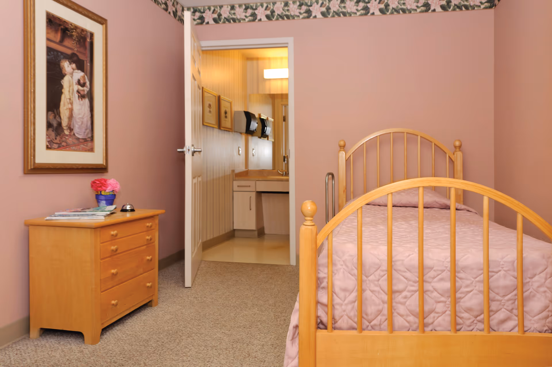 A bedroom with a wooden bed frame and a pink quilted bedspread. To the left is a wooden dresser with a small vase of pink flowers and some magazines on top. A framed painting hangs on the pink wall above the dresser. The door to the room is open, revealing a bathroom with a sink, mirror, and wall-mounted soap dispensers.