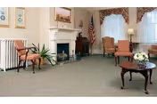 A cozy living room with a fireplace, two armchairs, a wooden coffee table with a flower arrangement, an American flag in the corner, and framed artwork on the walls. The room has carpeted flooring and large windows with curtains.