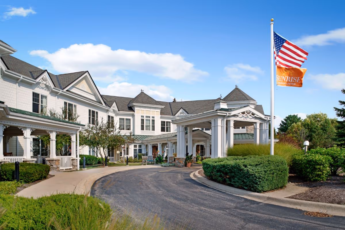 Front entrance of a large white senior living building with a covered porte-cochère, driveway, landscaping, and an American flag.