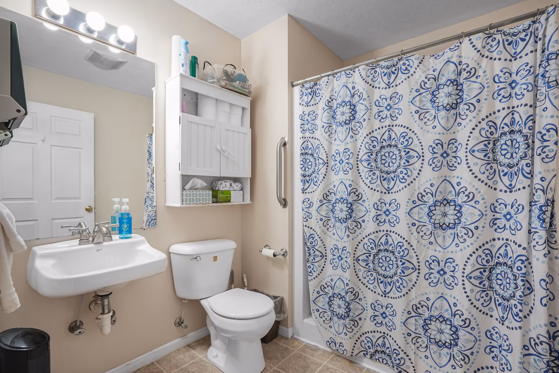A clean bathroom with a white sink and faucet, a toilet, and a bathtub with a blue and white patterned shower curtain. Above the toilet is a white wall cabinet stocked with toilet paper and other supplies. A mirror with lights is above the sink, and a towel hangs on the left side. The floor is tiled, and the walls are painted beige.
