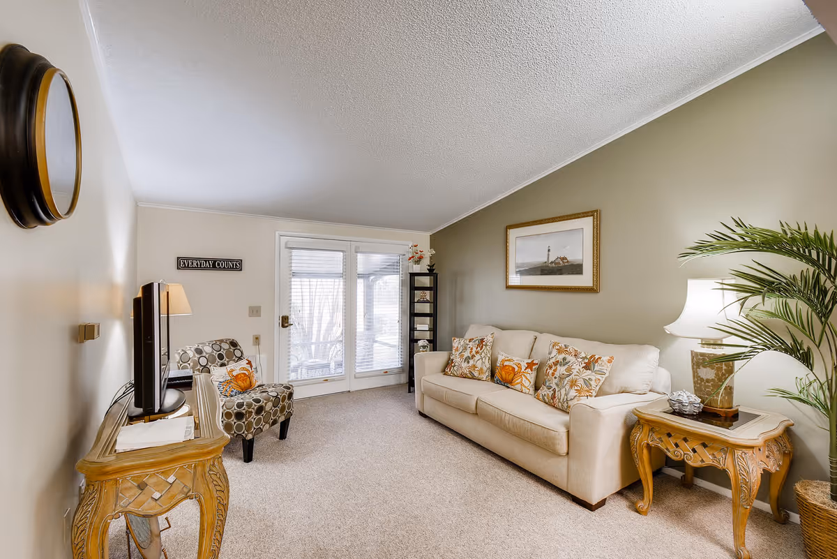 A cozy living room with a beige sofa adorned with floral and orange patterned cushions, a patterned armchair, a wooden side table with a lamp, a potted plant, a wall clock, and a framed picture of a lighthouse on a green accent wall. There is a glass door with blinds leading outside and a small black shelving unit with decorative items.