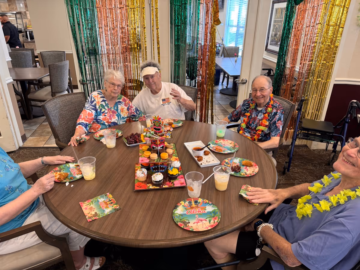 Several elderly residents sit around a decorated round table enjoying a tropical-themed party with snacks, cupcakes and drinks.