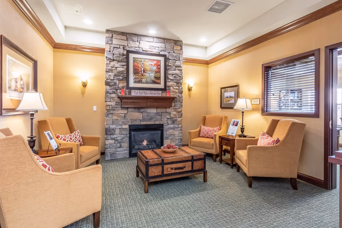 A cozy living room area with four beige armchairs arranged around a wooden coffee table designed like a trunk. The room features a stone fireplace with a framed painting above it, warm yellow walls, carpeted floor, and several lamps providing soft lighting. There are framed pictures on the walls and windows with blinds on the right side.