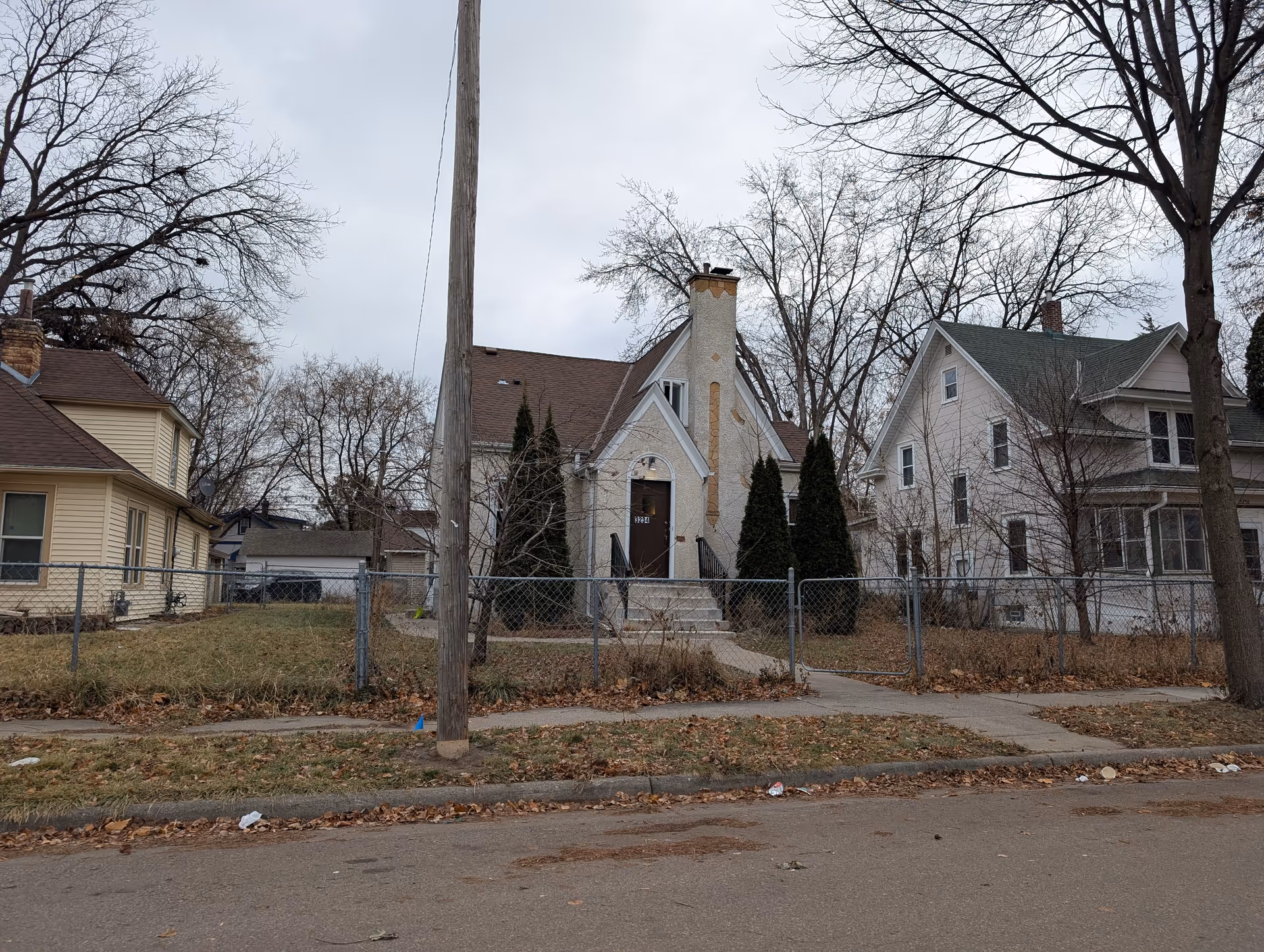 Front exterior of a small house with a chimney and front steps behind a chain-link fence, flanked by neighboring homes on a cloudy day.