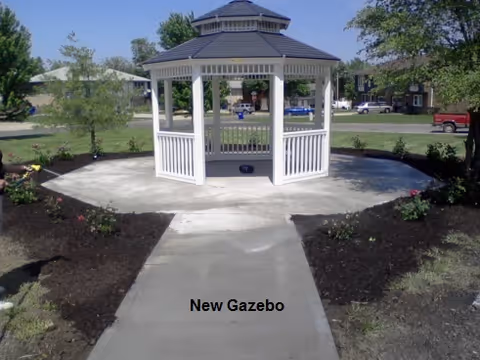 White gazebo at the end of a paved walkway surrounded by mulch beds and lawn with buildings in the background.