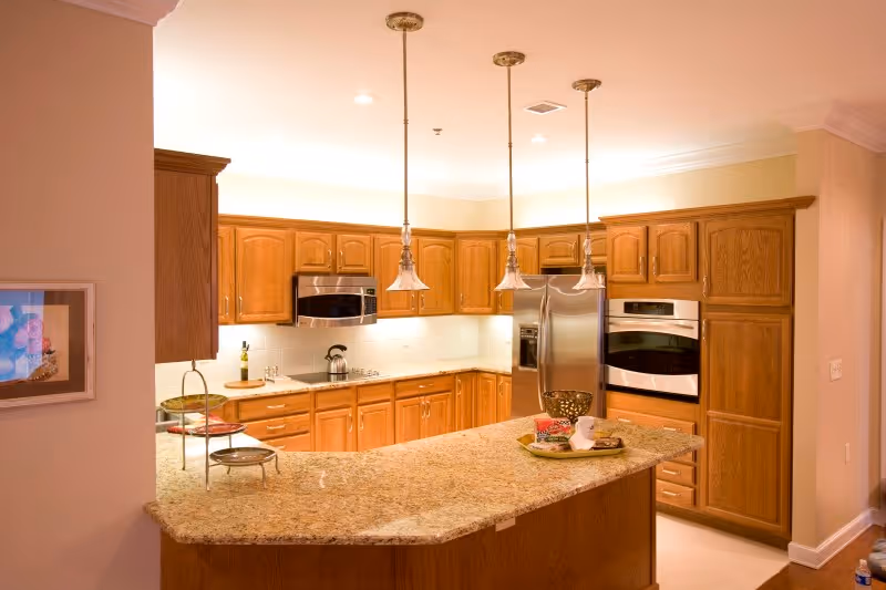 A spacious kitchen with wooden cabinets, granite countertops, and stainless steel appliances including a refrigerator, microwave, and built-in oven. Three pendant lights hang above the kitchen island, which has a tray with decorative items on it. The kitchen is well-lit with recessed lighting and under-cabinet lights.