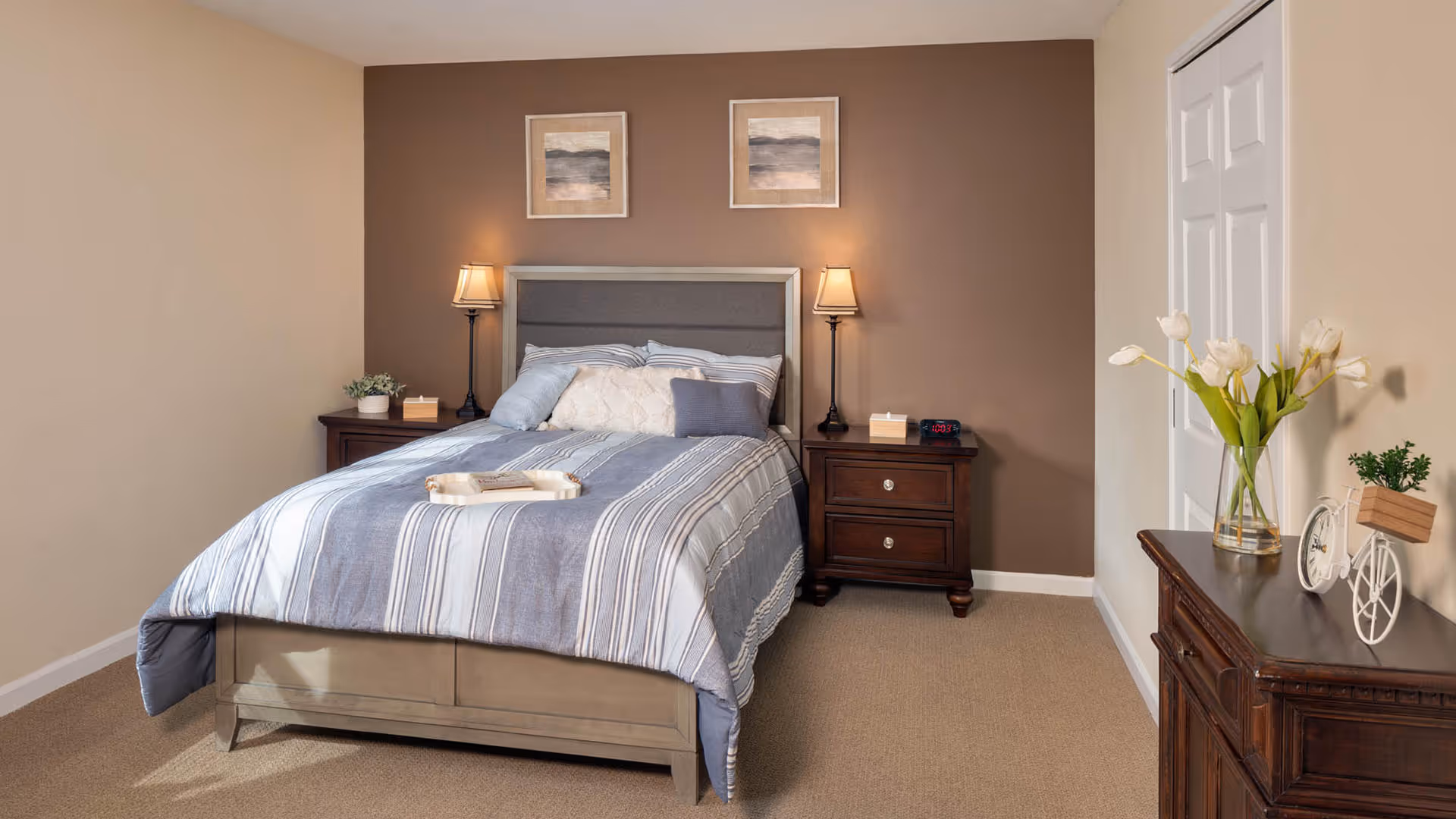 A neatly made bed with a striped blue and white comforter and multiple pillows in a bedroom. There are two dark wooden nightstands on either side of the bed, each with a lamp and small decorative items. The wall behind the bed is painted brown and has two framed pictures hanging above the headboard. To the right, there is a wooden dresser with a vase of white tulips and a decorative white bicycle. The room has beige carpet and light-colored walls.
