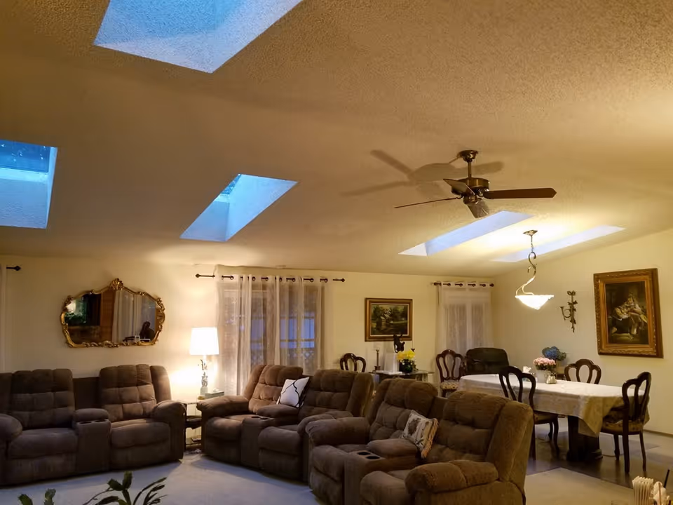 A cozy living and dining area with multiple brown recliner chairs arranged in a semi-circle, a dining table with six chairs covered with a white tablecloth, decorative paintings on the walls, sheer curtains on the windows, and skylights on the ceiling providing natural light. A ceiling fan and a hanging light fixture are also visible.