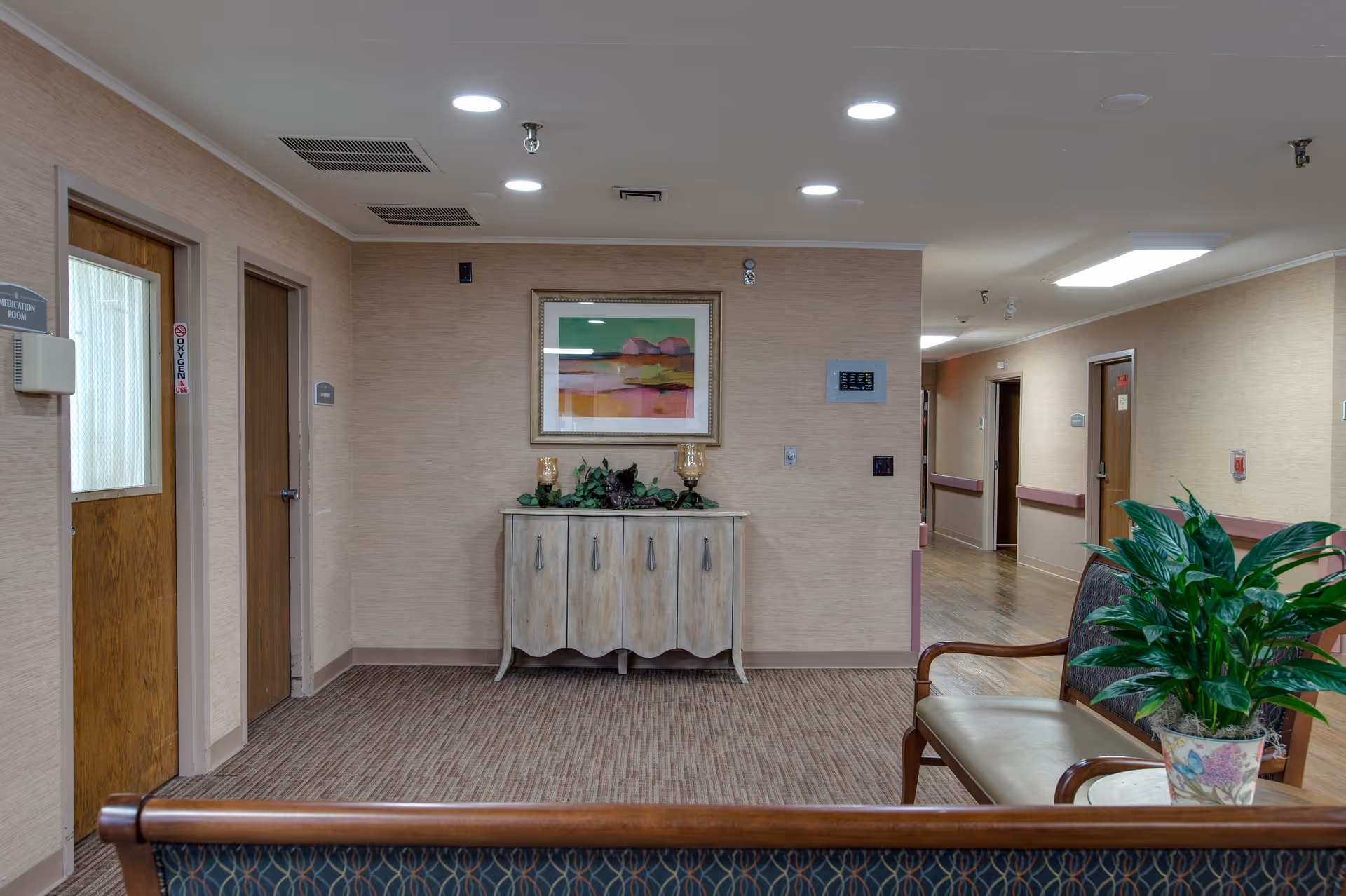 Interior hallway of a nursing facility with beige walls and carpeted floor. There are wooden doors on the left and right sides, a small wooden cabinet with decorative items and a framed abstract painting on the wall. In the foreground, there is a wooden bench with patterned upholstery and a potted plant on a small table beside it.