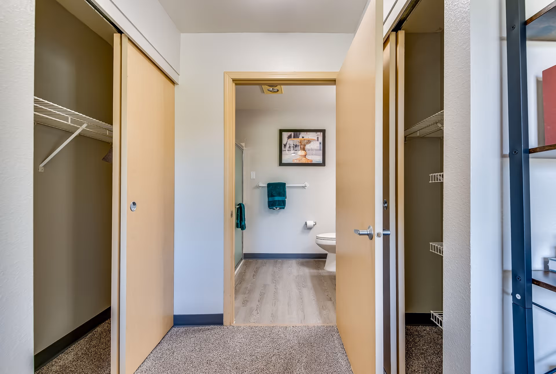 View from a carpeted room with two open closets on either side, looking through an open door into a bathroom with light wood flooring, a toilet, a glass shower door, teal towels hanging on a towel rack, and a framed picture on the wall.