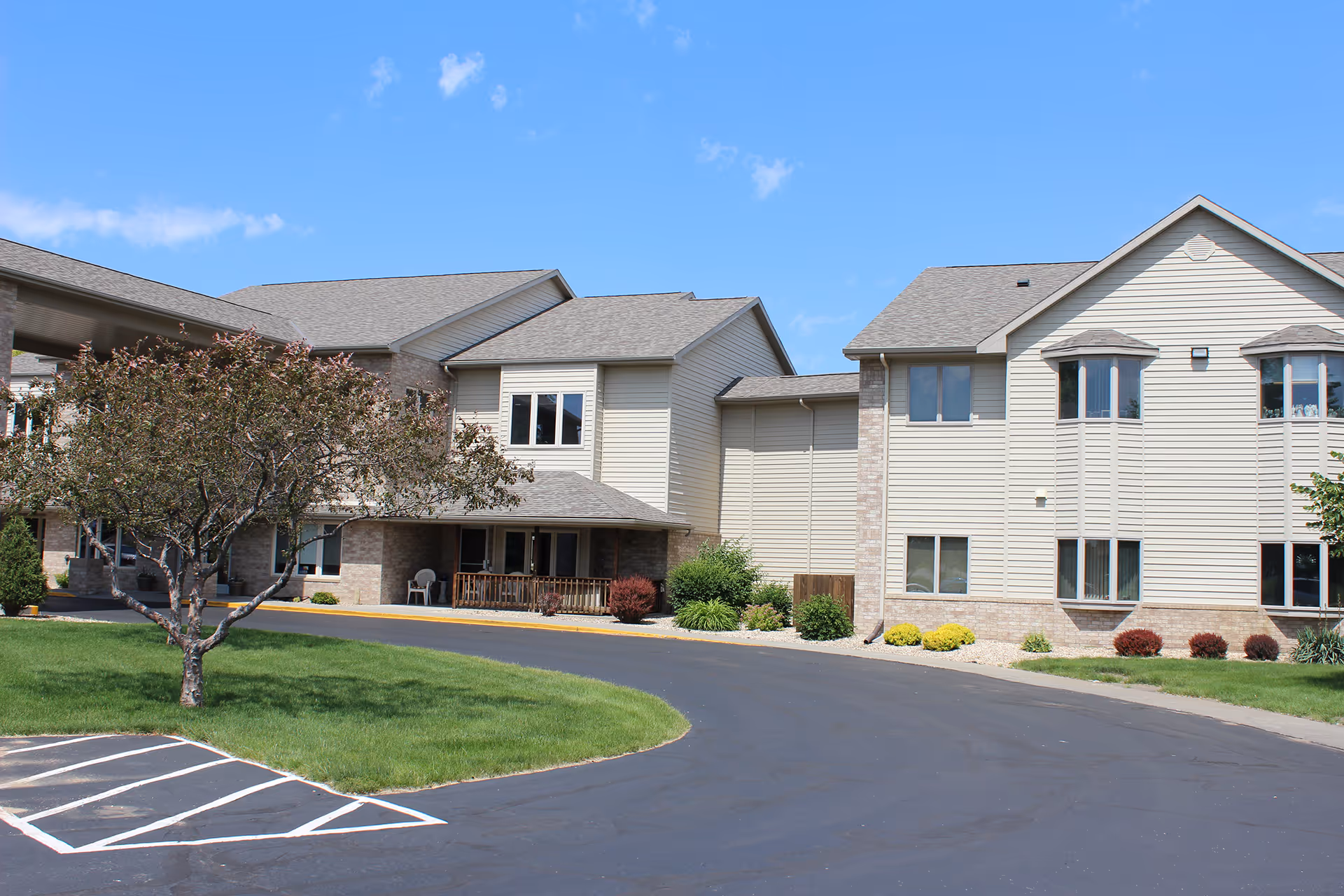 Exterior view of a senior living facility named Countryside Living with beige siding and brick accents, a curved driveway, green lawn, and a tree in front under a blue sky with a few clouds.