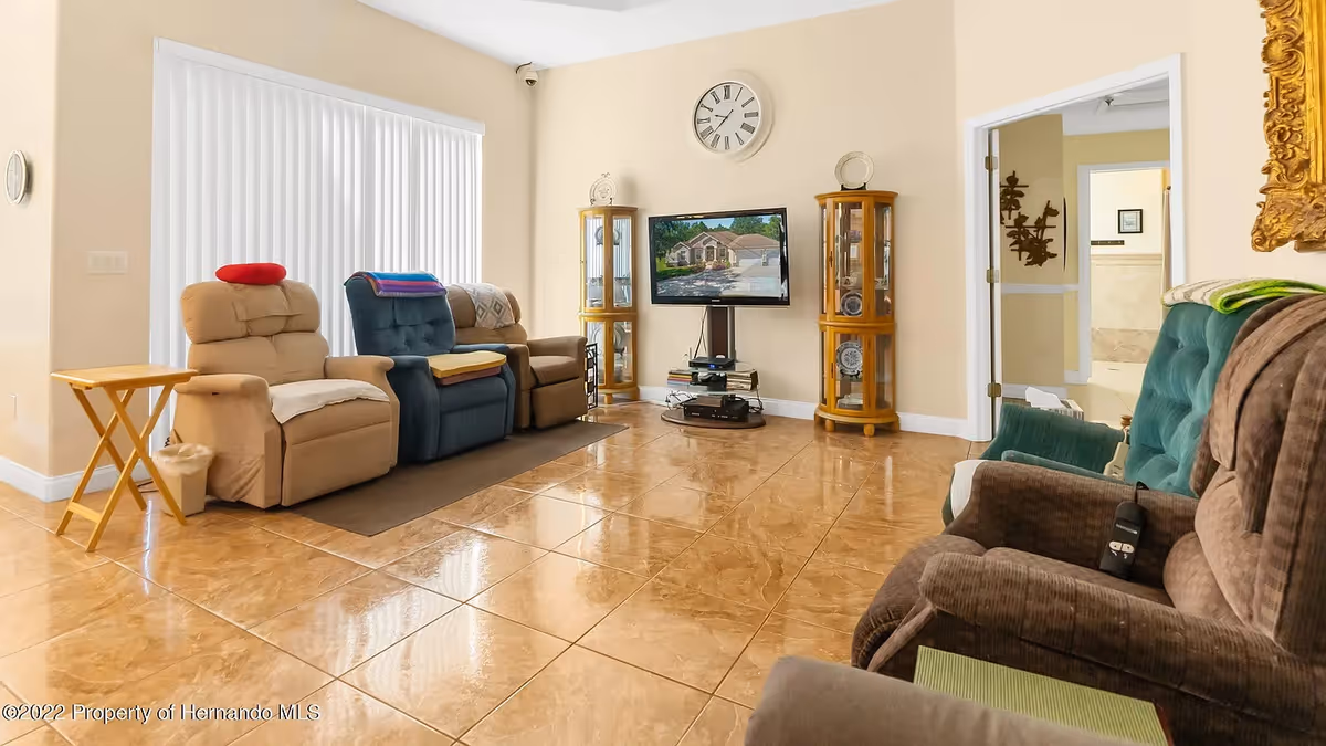 A cozy living room with several recliner chairs arranged around a flat-screen TV mounted on a stand. The room has beige walls, a large window with vertical blinds, two wooden display cabinets with glass doors, a wall clock above the TV, and a tiled floor. There is a small wooden side table next to one of the chairs and a doorway leading to another room.