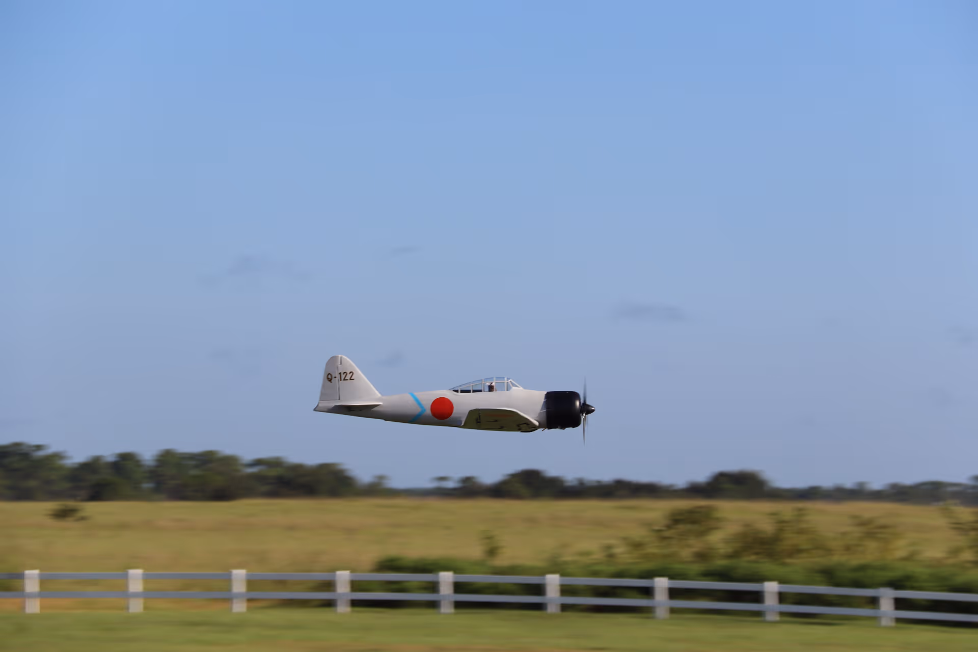 A vintage single-engine airplane flying low over a grassy field with a white fence under a clear blue sky.