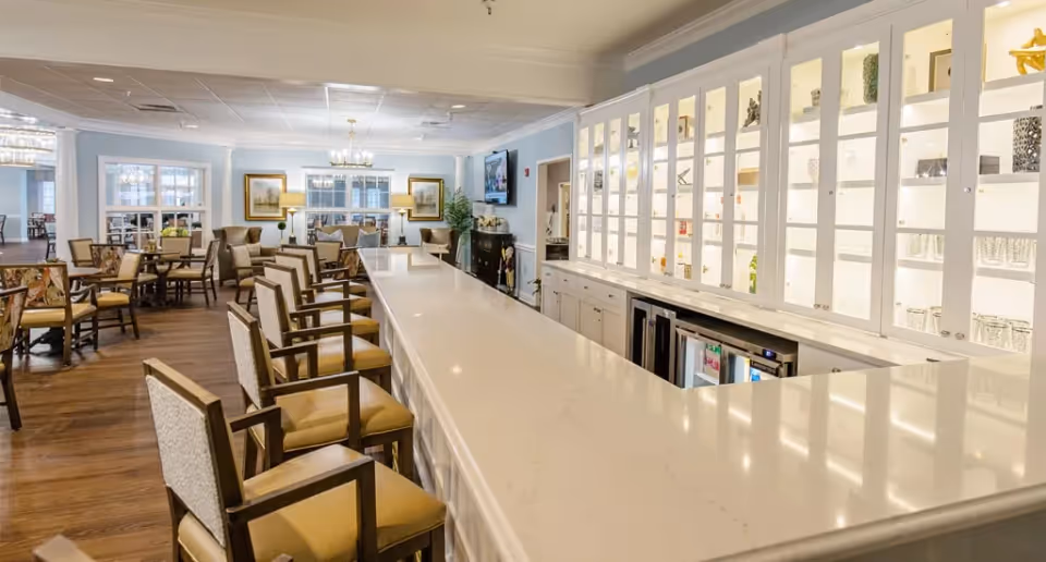 A spacious and well-lit dining area with a long white countertop bar lined with cushioned chairs. Behind the bar are glass cabinets displaying glassware and decorative items. The room features wooden flooring, multiple dining tables with chairs, and a cozy seating area with armchairs near the windows. The walls are painted light blue with white trim, and there are framed pictures and a television mounted on the wall.