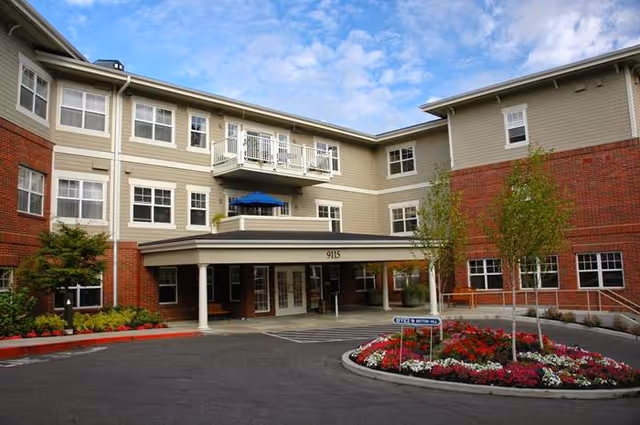 Exterior view of a three-story senior living facility building with beige siding and red brick accents. The entrance has a covered driveway with white columns and the number 9115 displayed above. There is a circular flower bed with colorful flowers and small trees in front of the entrance. The sky is partly cloudy.