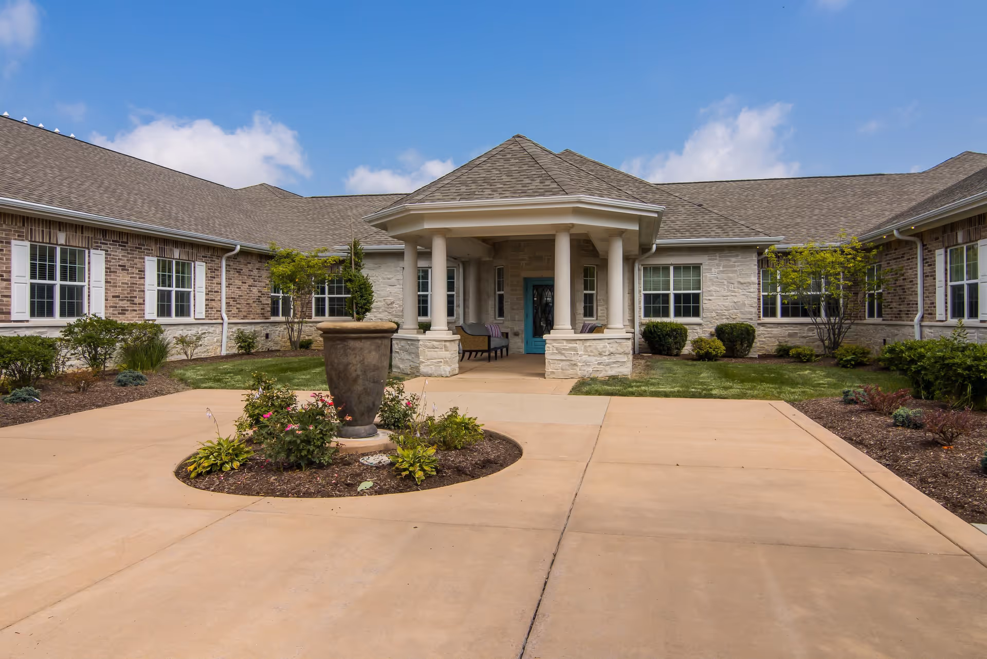 Front entrance of a single-story memory care building with a covered portico, columns, circular driveway, and landscaped planters.
