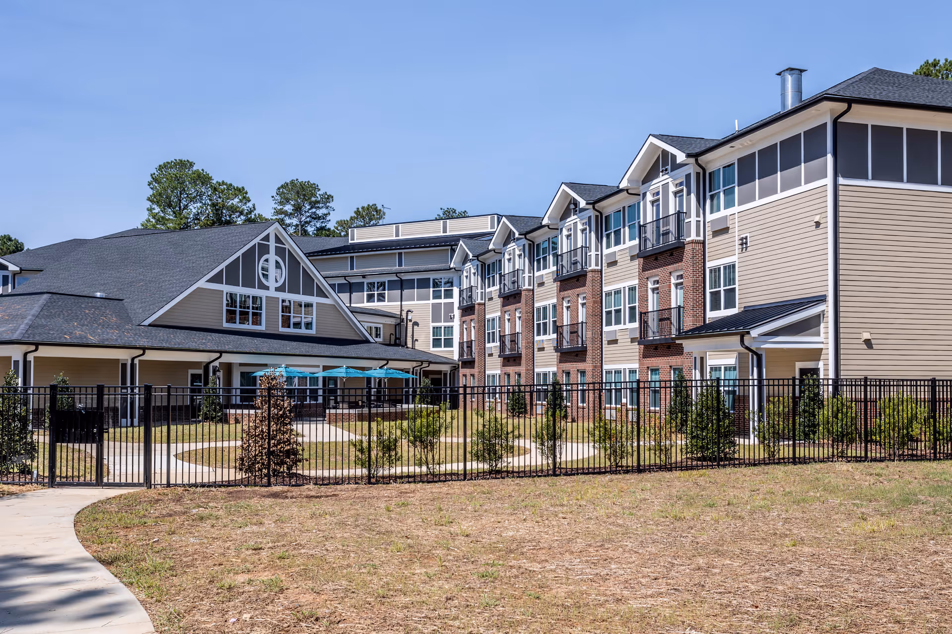 Exterior view of a multi-story senior living building with a fenced courtyard and patio umbrellas.
