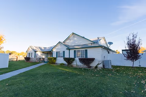 Single-story residential building with white siding and green shutters, surrounded by a well-maintained lawn and a white fence under a clear blue sky.