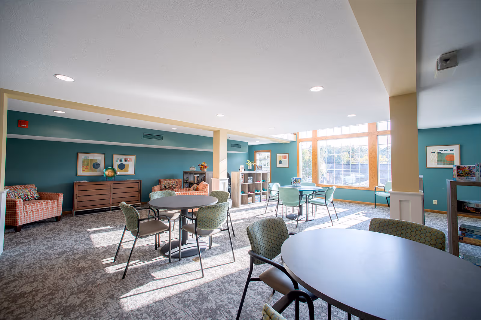A bright and spacious common area with large windows letting in natural light. The room features several round tables with green patterned chairs, two orange patterned armchairs, a wooden sideboard, and shelves with books and board games. The walls are painted teal with framed abstract art hanging on them.