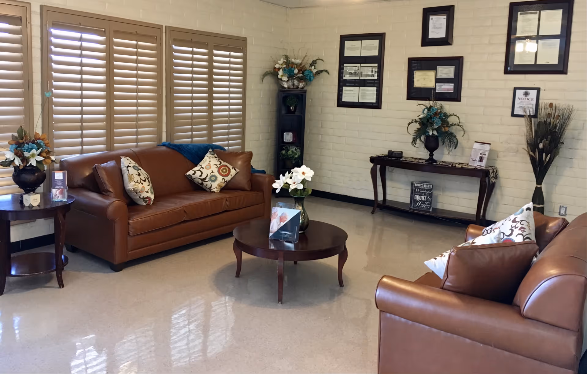 A cozy sitting area with two brown leather sofas adorned with decorative pillows. A round wooden coffee table with a flower vase is placed in the center. The room has beige walls with framed certificates and decorations, a tall black shelf with plants and flowers, and a wooden side table with a flower arrangement. Large windows with wooden shutters allow natural light to enter the space.