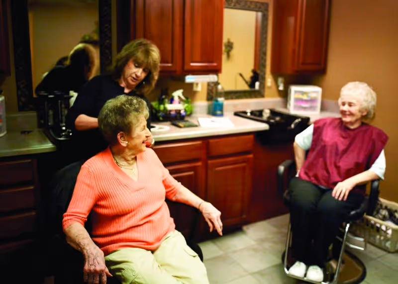 A hairdresser styles the hair of an elderly woman seated in a chair, while another elderly woman wearing a maroon salon cape sits nearby in a salon setting with wooden cabinets and a mirror on the wall.