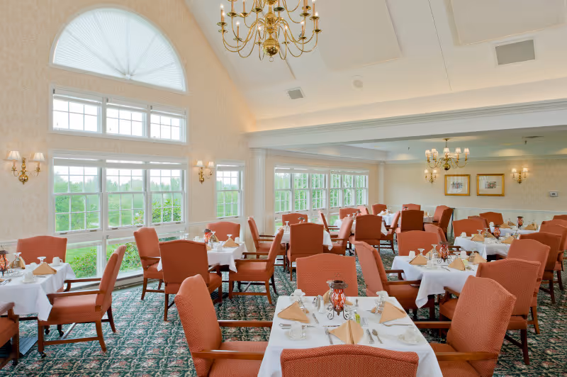 A bright and elegant dining room with multiple tables covered in white tablecloths, each set with beige napkins, plates, cups, and silverware. The room features large windows allowing natural light to fill the space, red upholstered chairs, chandeliers hanging from a high ceiling, and patterned carpet flooring.