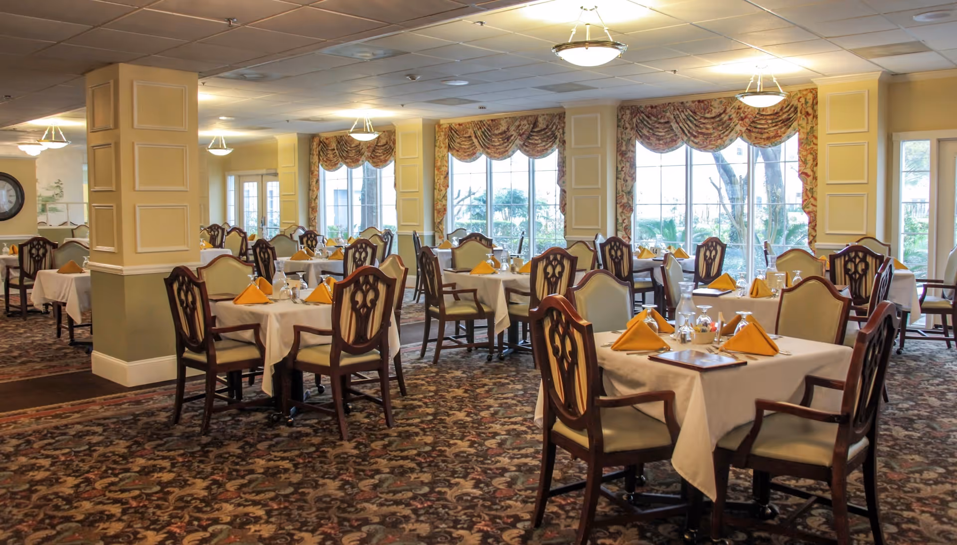 A spacious dining room with multiple tables covered in white tablecloths, each set with yellow folded napkins, glassware, and silverware. The room features large windows with floral valances allowing natural light to fill the space, patterned carpet flooring, and elegant wooden chairs with cushioned seats.