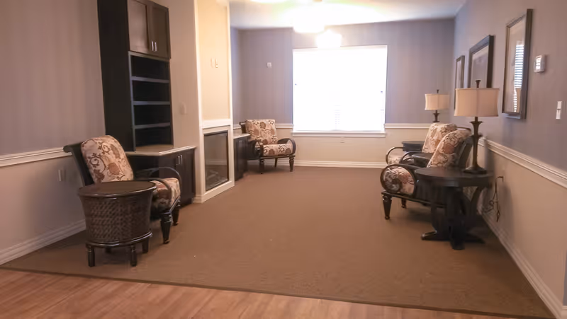 A quiet senior living common area with four upholstered armchairs arranged along the walls, two side tables with lamps, a built-in shelving unit, and a window letting in natural light.