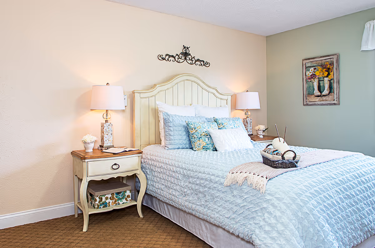 A cozy bedroom with a cream-colored wooden bed frame and a light blue textured bedspread. The bed is adorned with multiple decorative pillows in shades of blue and white. On either side of the bed are matching nightstands with table lamps, a small flower vase, and decorative items. The walls are painted in soft beige and green tones, with a framed floral painting hanging on the green wall.