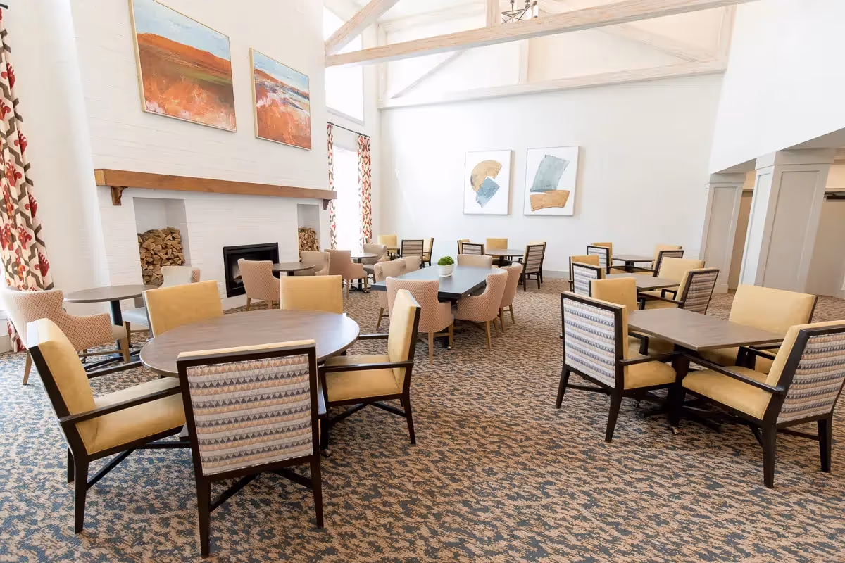 A bright and spacious dining room with multiple round and rectangular tables surrounded by cushioned chairs. The room features a high ceiling with exposed beams, large windows with patterned curtains, a fireplace with stacked firewood on either side, and abstract artwork on the walls.