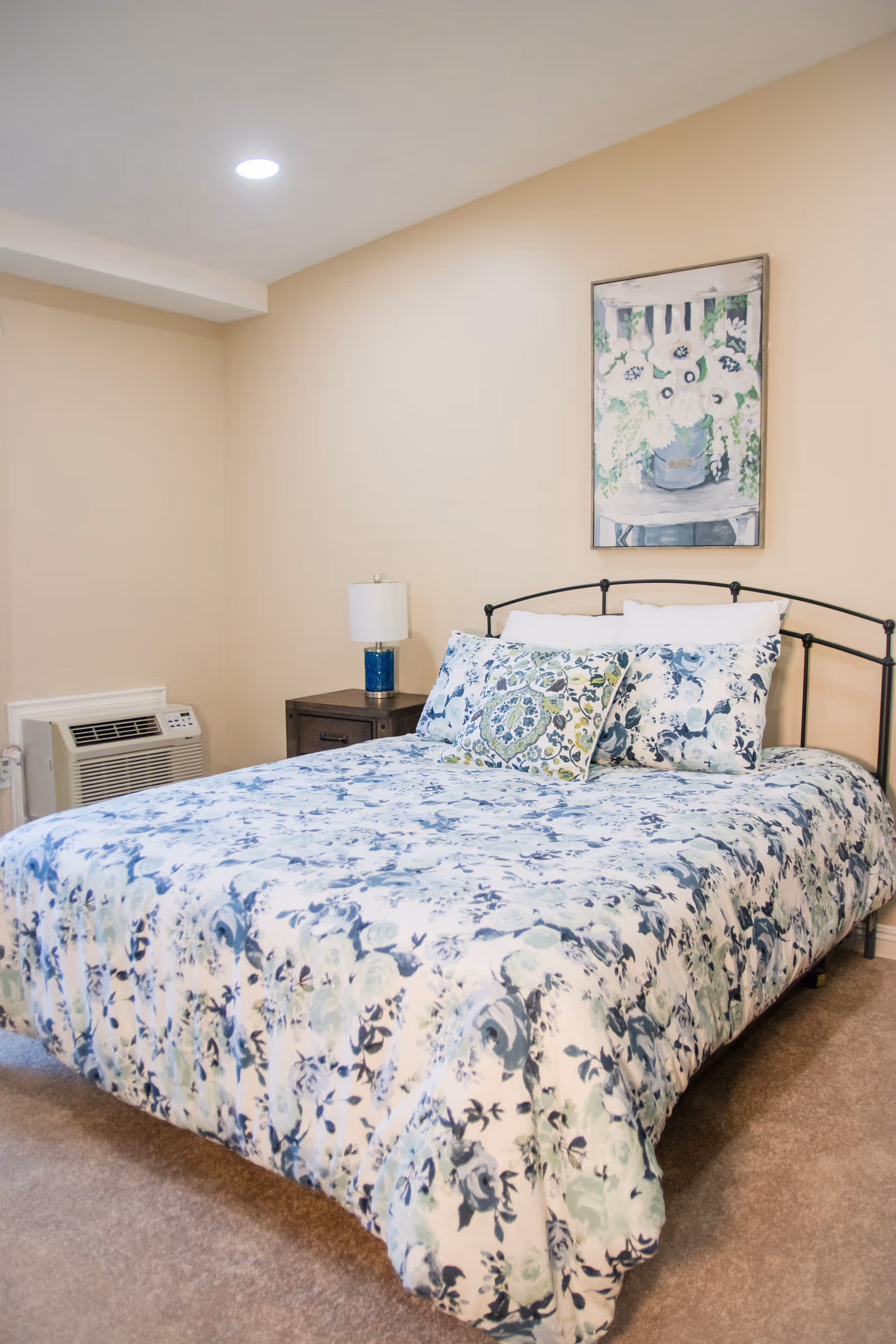 A bedroom with a bed featuring a floral patterned comforter and pillows in shades of blue and green. There is a dark metal headboard, a wooden nightstand with a blue lamp, and a framed floral painting on the beige wall above the bed. The room has beige walls and carpeted floor, with a wall-mounted air conditioning unit visible on the left side.