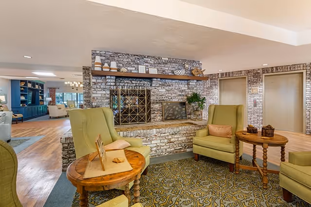 A cozy seating area in a senior living facility featuring three green upholstered armchairs arranged around two wooden side tables. The background includes a decorative brick wall with a built-in shelf holding various decorative items and two elevator doors. The floor is covered with a patterned carpet and wood flooring.