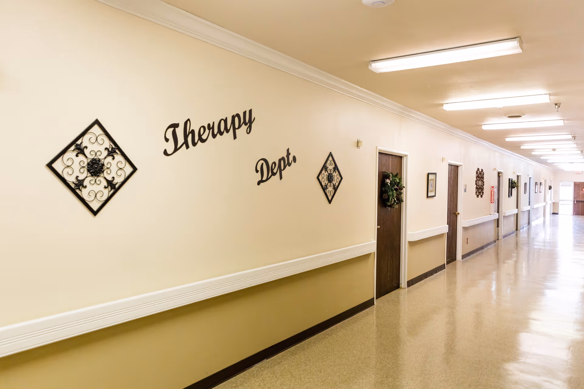 A long, well-lit hallway in a healthcare facility with beige walls and a polished floor. The wall on the left has decorative metal wall art and the words 'Therapy Dept.' mounted on it. Several closed wooden doors line the hallway, some adorned with wreaths. Handrails run along both sides of the corridor.