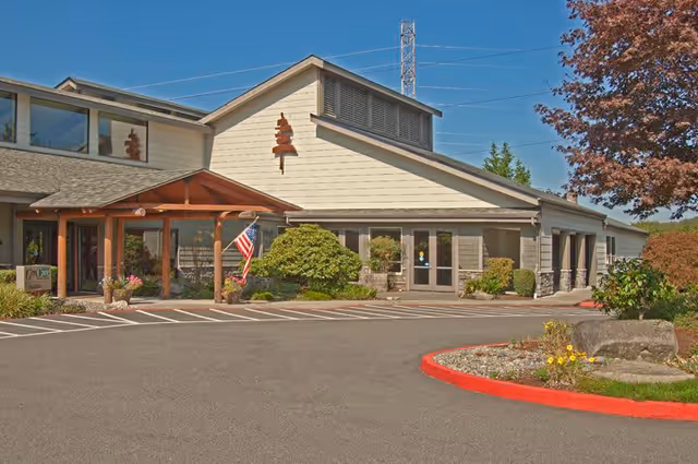 Exterior view of a single-story building with beige siding and a sloped roof, featuring a covered entrance with wooden beams and an American flag. There are bushes and landscaping around the building, a parking area in front, and a tree with red leaves on the right side.