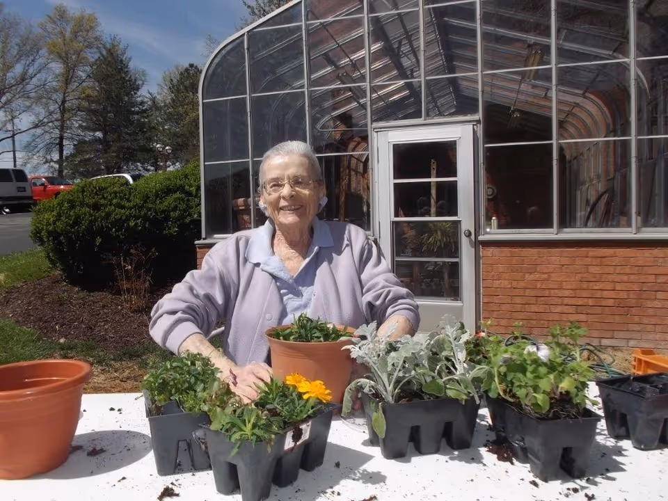 An elderly woman wearing glasses and a light purple cardigan is sitting at a table outdoors with several small potted plants and seedlings in front of her. Behind her is a greenhouse with a glass door and brick base. The sky is clear and trees and bushes are visible in the background.