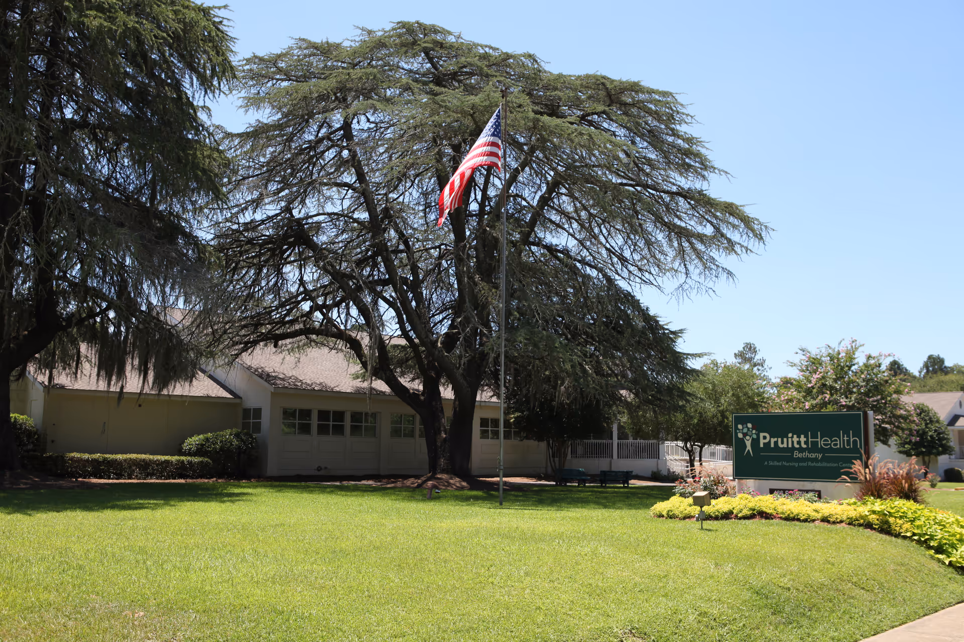 Front lawn and entrance of PruittHealth - Bethany with an American flag on a flagpole, large trees, and the facility sign.